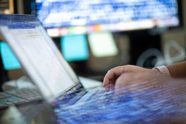 A technician works with a laptop in front of a large screen displaying code.